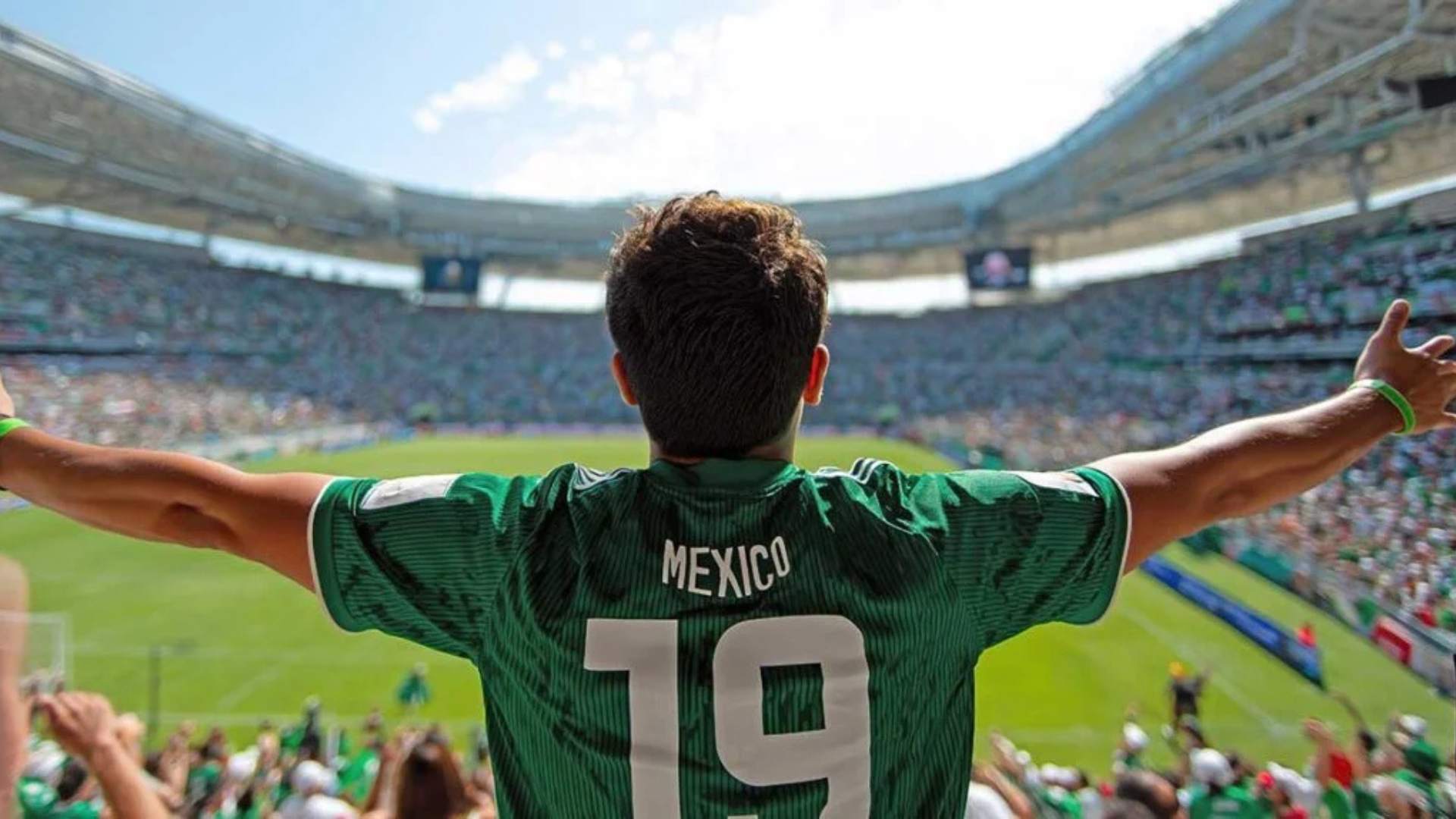 Soccer player in a green Mexico jersey with arms raised, celebrating in a packed stadium.
